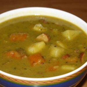 A bowl of traditional German pea soup garnished with carrots, potatoes, and sausage pieces.