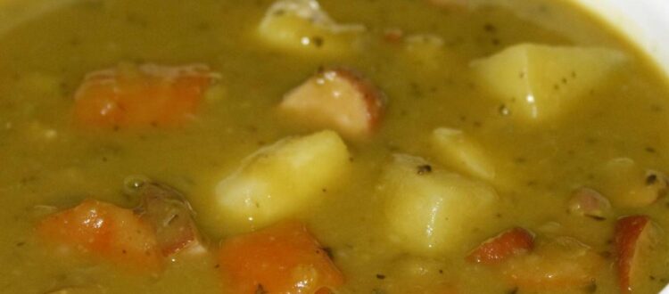 A bowl of traditional German pea soup garnished with carrots, potatoes, and sausage pieces.