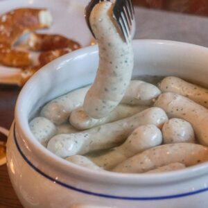 A fork lifting a Weisswurst sausage from a bowl filled with several Weisswurst sausages in hot water, with pretzels in the background.