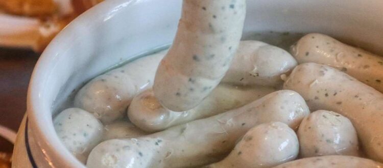 A fork lifting a Weisswurst sausage from a bowl filled with several Weisswurst sausages in hot water, with pretzels in the background.